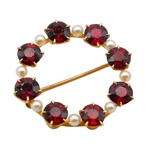 May include: A gold-toned brooch featuring a circular design. The brooch is adorned with alternating round, deep red gemstones and small, round pearls. A gold pin secures the brooch.
