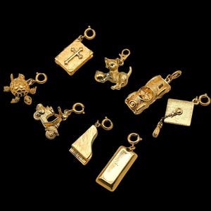 May include: A collection of eight gold-toned metal charms featuring various shapes including a book with a cross, a cat, a turtle, a car, a scooter, a grand piano, a graduation cap with a tassel, and a rectangular bar, each with a jump ring.