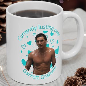Puede incluir: Una taza de cerámica blanca con un gráfico de un hombre sin camisa y el texto en color turquesa "Currently lusting over Garrett Graham" rodeado de pequeños iconos de corazones turquesas. La taza tiene un asa y acabado brillante.