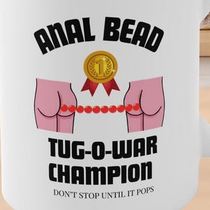 Puede incluir: Taza de cerámica blanca con el texto "ANAL BEAD TUG-O-WAR CHAMPION" y un gráfico de dos nalgas rosas conectadas por una cadena de cuentas. La taza también presenta un gráfico de medalla de oro.