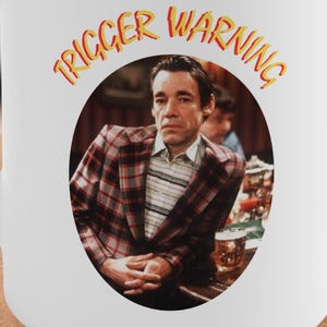 Può includere: Tazza in ceramica bianca con manico. La tazza presenta un'immagine circolare di un uomo con una giacca a quadri con le parole "TRIGGER WARNING" in giallo e arancione. La tazza è su una superficie di legno.