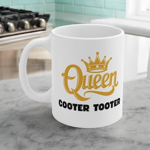 May include: White ceramic mug with a large handle. The mug features the word "Queen" in gold script, topped with a gold crown, and the words "COOTER TOOTER" in black block letters. The mug is on a light gray surface.
