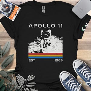 May include: Black t-shirt with a graphic of an astronaut on the moon. The text "APOLLO 11" is above the graphic, and "EST. 1969" is below. The design includes a red, yellow, and blue horizontal stripe.