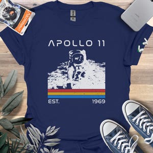 May include: A navy blue t-shirt with a vintage-style graphic of an astronaut on the moon. The graphic includes the text "Apollo 11", "Est." and "1969".