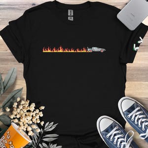 Pode incluir: Camiseta preta com um gráfico de um carro DeLorean passando por chamas. O carro é branco com rodas vermelhas. As chamas são laranja e amarelas. A camiseta é de gola redonda.