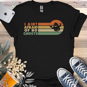 May include: Black t-shirt with the text "I AINT AFRAID OF NO GHOSTS" in orange, yellow, and green. A retro-style graphic of a ghost is on the right side of the text. The shirt is paired with blue sneakers.