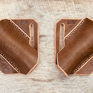 May include: Two brown leather holsters with a stitched design and the text "L-45" and "R-45". Each holster has a rectangular base with slots and a curved pocket. The holsters are designed for a firearm.