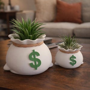 May include: Two white ceramic money bag planters with green dollar signs. The larger planter has a green plant, and the smaller one has a smaller green plant. Both planters have a rope-like detail around the top.