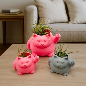 May include: Three cat-shaped planters in pink and gray, each holding a green plant. The planters are on a wooden table. The cats are giving the middle finger. The background includes a couch and side table.