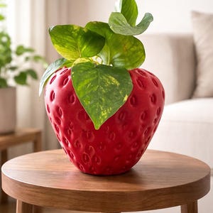 Puede incluir: Una maceta roja con forma de fresa con una superficie texturizada, llena de hojas verdes y moteadas de amarillo. La maceta está sobre una mesa redonda de madera. El fondo incluye un sofá blanco y otra planta.
