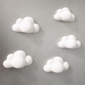 Puede incluir: Un conjunto de cinco imanes blancos con forma de nube en tres dimensiones. Cada imán presenta un diseño redondeado y esponjoso con una superficie texturizada, dispuestos de forma dispersa sobre una superficie metálica de color gris.