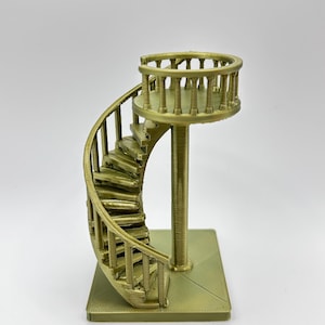 May include: A gold-colored miniature spiral staircase with a circular platform at the top. The staircase has detailed steps and a railing, supported by a central column and a square base. The design is intricate and decorative.