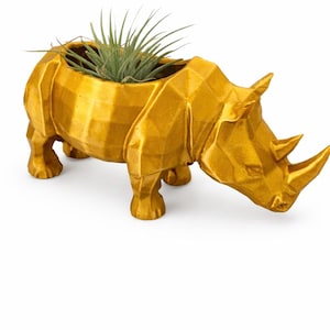 May include: A gold-colored, geometric rhino planter with a small air plant inside. The rhino is in a walking position, with its head lowered. The planter is made of a hard material and has a textured surface.