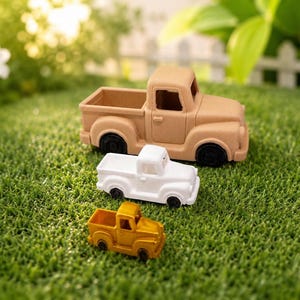 May include: Three toy pickup trucks in varying sizes and colors are arranged on a bed of green grass. The largest truck is tan, the medium truck is white, and the smallest truck is yellow. The trucks have black wheels.