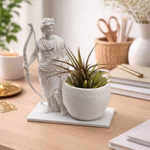 Puede incluir: Una estatua blanca de un hombre con arco y flecha se encuentra junto a una maceta blanca con un diseño geométrico. La maceta contiene una planta verde con hojas largas y finas. Los objetos están sobre una superficie de madera.