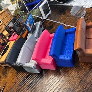 May include: A collection of miniature furniture pieces, including sofas in pink, blue, and brown, and small televisions with screens displaying images. The items are arranged on a wooden surface, showcasing a variety of colors and textures.