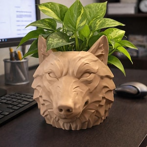 Peut inclure: Un pot de fleur en forme de loup de couleur beige, contenant une plante verte à feuilles. Le pot présente des détails, notamment les oreilles, les yeux et le museau du loup. La plante a des feuilles vertes avec des accents jaunes.