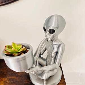 May include: A silver-colored alien figurine holding a small, round planter with a succulent plant. The alien is seated with its legs crossed and is resting on a circular base. The planter is also silver.