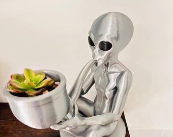 Puede incluir: Una figura alienígena de color plateado sentada con las piernas cruzadas, sosteniendo una maceta cilíndrica en sus manos. El alienígena tiene grandes ojos negros y un acabado metálico liso. Una pequeña planta suculenta verde y roja está dentro.
