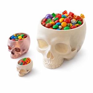 Puede incluir: Tres cuencos con forma de calavera llenos de caramelos de colores. La calavera más grande es blanquecina y contiene una variedad de caramelos. Una calavera más pequeña de color cobre y otra más pequeña blanquecina también están llenas de caramelos. Ideal para Halloween.