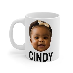 Op de afbeelding: Witte keramische mok met een foto van een babymeisje met een strik op haar hoofd. De mok heeft de naam "CINDY" in zwarte letters onder de foto.