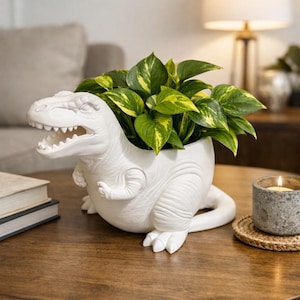 May include: A white ceramic dinosaur planter, filled with green and yellow leafy plants, sits on a wooden table. The planter is a detailed sculpture of a Tyrannosaurus Rex. A candle and books are also on the table.