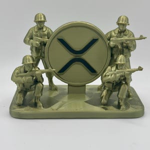 Puede incluir: Escultura dorada que presenta cuatro figuras de soldados que rodean un gran emblema con un símbolo negro en forma de X. Los soldados están en poses de combate, sosteniendo rifles. La base es rectangular y tiene un tema militar.