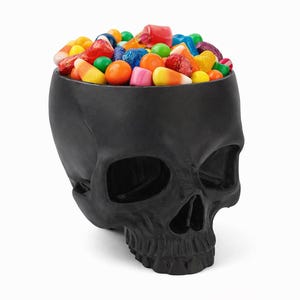 Pu&ograve; includere: Una ciotola nera a forma di teschio piena di caramelle colorate. Le caramelle includono dolci arancioni, gialli, rossi, verdi e blu. La ciotola &egrave; in materiale nero opaco, su sfondo bianco.