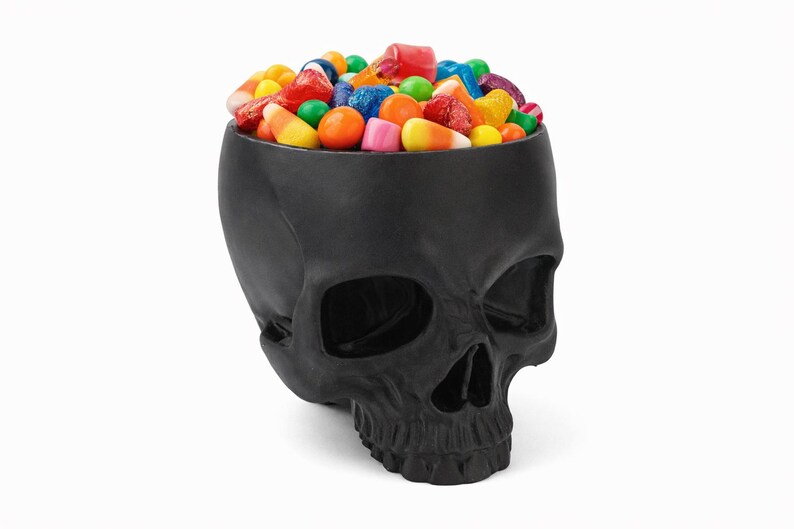 Pu&ograve; includere: Una ciotola nera a forma di teschio piena di caramelle colorate. Le caramelle includono dolci arancioni, gialli, rossi, verdi e blu. La ciotola &egrave; in materiale nero opaco, su sfondo bianco.