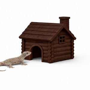 Op de afbeelding: Een bruin reptielenhuis in blokhutstijl met een schoorsteen en een klein raam. Een lichtgekleurde gekko staat bij de ingang. De reptielenhabitat is ontworpen om op een miniatuurhut te lijken.