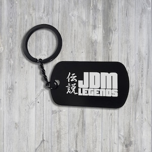 Puede incluir: Llavero de metal negro con un logotipo JDM LEGENDS plateado y caracteres japoneses. El llavero está unido a una cadena plateada.