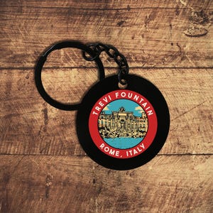 May include: Black round keychain featuring the Trevi Fountain in Rome, Italy. The image is set within a red circle, with the words "Trevi Fountain" and "Rome, Italy" in white text. The keychain has a black ring and chain.