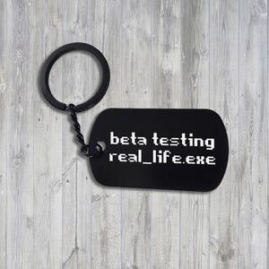Könnte beinhalten: Schwarzer Schlüsselanhänger aus Metall mit einem schwarzen Anhänger, auf dem "beta testing real_life.exe" steht.