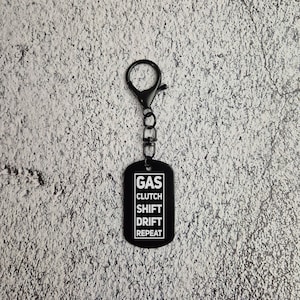 May include: Black metal keychain with a silver key ring. The keychain has a black rectangular tag with white text that reads "GAS CLUTCH SHIFT DRIFT REPEAT".