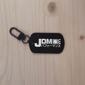 Può includere: Portachiavi nero con un anello in metallo argentato. Il portachiavi ha un'etichetta rettangolare nera con testo bianco che dice "JDM" e "パフォーマンス" in giapponese.