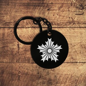 Könnte beinhalten: Schwarzer runder Schlüsselanhänger mit einem weißen, stilisierten floralen Design. Der Schlüsselanhänger hat einen schwarzen Ring und eine Kette. Das Design ist ein weißes Sonnenstrahlenmuster auf schwarzem Hintergrund. Er liegt auf einer Holzoberfläche.