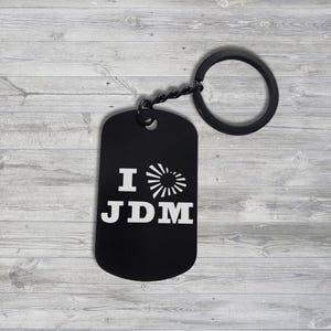 Könnte beinhalten: Schwarzer Schlüsselanhänger in Form einer Erkennungsmarke mit dem weißen Text "I JDM". Das "I" wird von einem stilisierten Sonnenmuster gefolgt. Der Schlüsselanhänger hat einen schwarzen Ring und eine kleine Kette. Der Hintergrund ist hellgraues Holzmaserung.