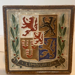 May include: A ceramic tile with a coat of arms featuring four lions in different colors and a banner with the word "LIMBURG". The tile is surrounded by a green leafy border with red berries.