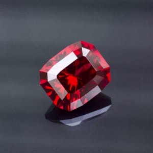 Może przedstawiać: Fasetowany, prostokątny, głęboko czerwony kamień szlachetny o wspaniałym połysku. Fasetki kamienia odbijają światło, tworząc olśniewający pokaz koloru i blasku. Kamień szlachetny jest osadzony na ciemnym tle, podkreślając jego bogaty kolor.