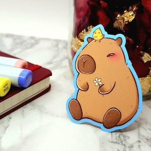 May include: A brown capybara sticker with a yellow bird on its head. The capybara is holding a white flower and has a light blue outline.