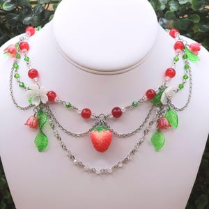 May include: A silver-toned necklace with a strawberry pendant. The necklace features red beads, green beads, white flowers, and dangling chains. The strawberry pendant is red and yellow. The necklace is displayed on a white mannequin.