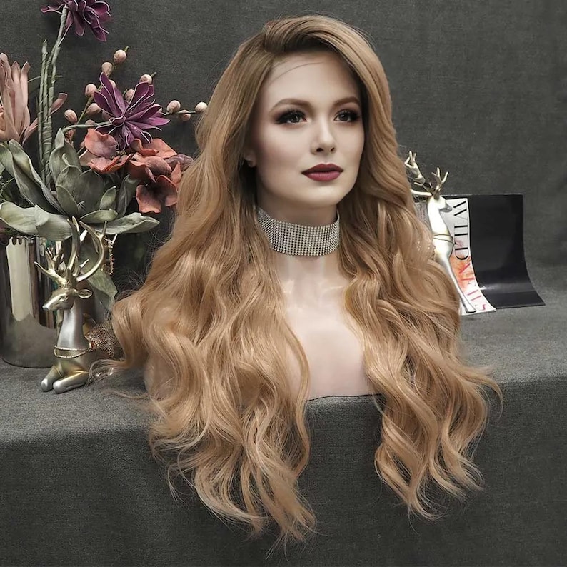 May include: A long, wavy blonde wig styled on a mannequin head. The wig features a natural-looking hairline and a layered cut. The mannequin is wearing a sparkling choker necklace. The background includes floral arrangements and decorative elements.