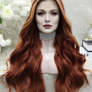 May include: A long, wavy wig with auburn and copper tones. The wig features a highlighted top and a natural-looking hairline. The hair is styled with loose waves, creating a voluminous and glamorous look. The wig is displayed on a mannequin head.