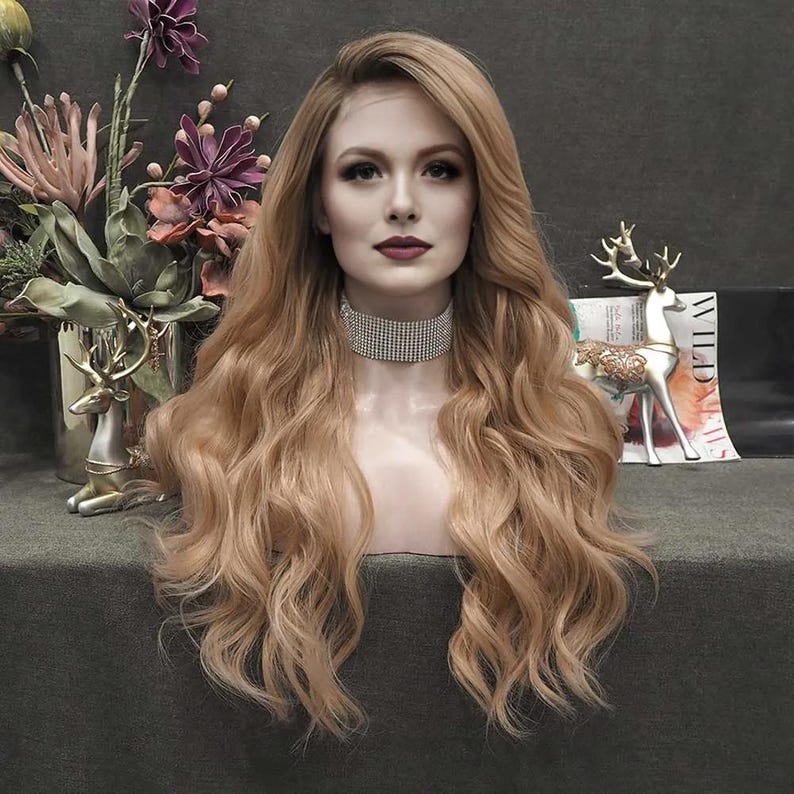 May include: A long, wavy, light brown wig on a mannequin head. The wig has a natural-looking hairline and is styled with loose curls. The mannequin is wearing a sparkling rhinestone choker. Decorative flowers and a deer figurine are in the background.