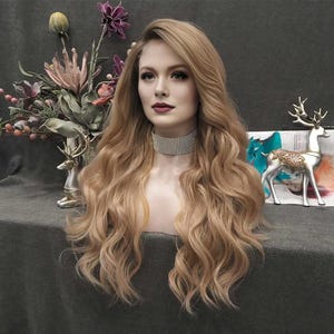 May include: A long, wavy blonde wig styled on a mannequin head. The wig features a natural-looking hairline and a soft, flowing texture. The mannequin is wearing a sparkling choker necklace. Decorative elements include flowers and deer figurines.