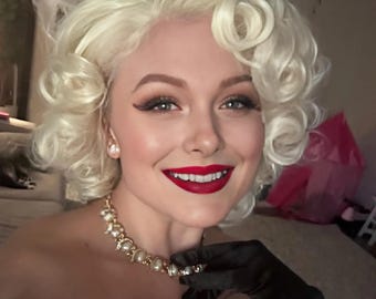 May include: A woman with blonde, curly hair, red lipstick, and a pearl necklace. She is wearing black gloves and has a white fur-like trim. The image is a close-up, showing her smiling face and makeup.