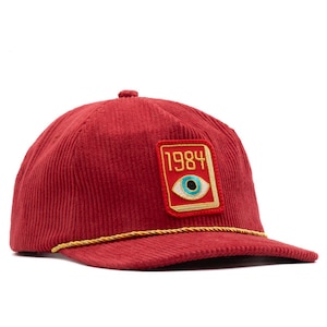 May include: A red corduroy baseball cap with a flat brim and a gold rope detail. The cap features a rectangular patch with the year "1984" and an eye design. The cap is a stylish accessory.