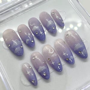 以下が含まれることがあります： 薄いパープルから濃いパープルへのグラデーションデザインの10本の付け爪のセット。各爪には、白い三日月と雲のデザインと、シルバーのグリッターのアクセントが施されています。