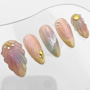 Può includere: Cinque unghie finte iridescenti rosa e bianche con accenti dorati. Le unghie sono decorate con conchiglie, perle e una stella marina.