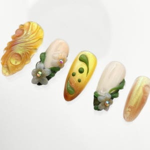 May include: Five press-on nails featuring a mix of cream and iridescent yellow finishes. Designs include raised 3D floral motifs with small embedded crystals, green leaf accents, and swirling yellow textures with raised dots on a smooth surface.